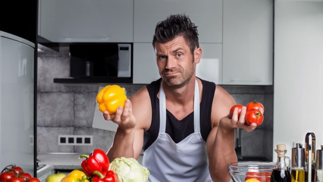 Strong healthy muscle sporty looking handsome charismatic man bodybuilder in black tank top and white apron leaning on kitchen counter with colorful vegetables, bottle of vinegar and spices on it.