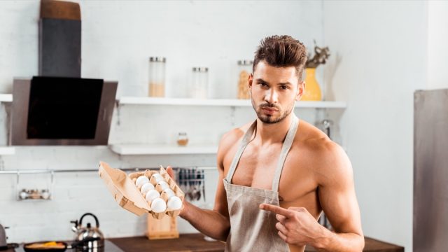 Bare-chested man in apron pointing with finger at egg carton.