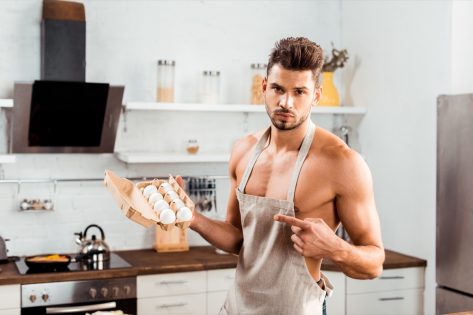 Bare-chested man in apron pointing with finger at egg carton.