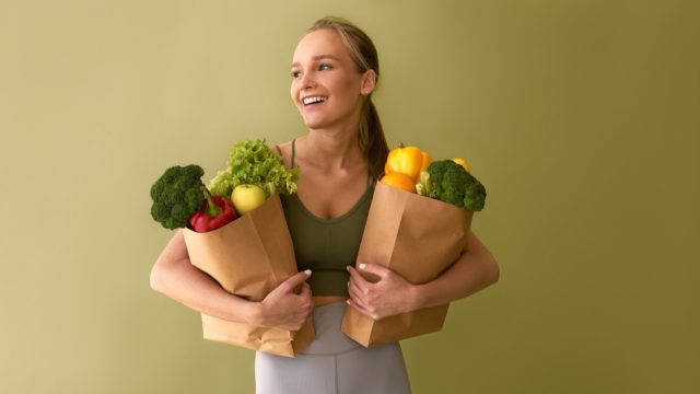 Attractive young woman holding bags of vegetables on a green background.