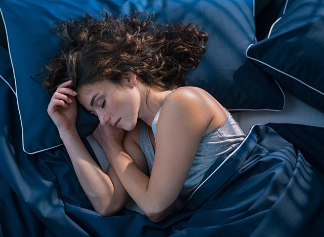 Top view of young woman sleeping on side in her bed at night. Beautiful girl sleeping profoundly and dreaming at home with blue blanket. High angle view of woman asleep with closed eyes.
