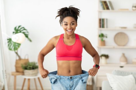 Happy African Woman After Slimming Showing Her Old Oversize Pants Standing Indoors, Smiling To Camera