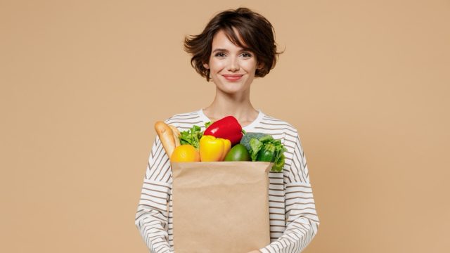 Young smiling caucasian cheerful happy fun vegetarian woman 20s in casual clothes hold paper bag with vegetables after shopping look camera isolated on plain pastel beige background studio portrait.