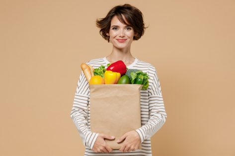 Young smiling caucasian cheerful happy fun vegetarian woman 20s in casual clothes hold paper bag with vegetables after shopping look camera isolated on plain pastel beige background studio portrait.