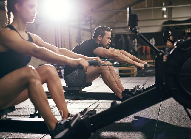 Side view of man and woman doing exercises with rowing machine at gym.