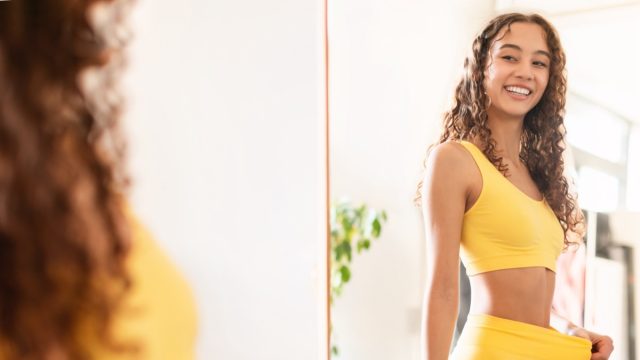 Happy fit woman looking at reflection in mirror and pulling fitness pants waist showing result of dieting and slimming, motivated after workout training at home.