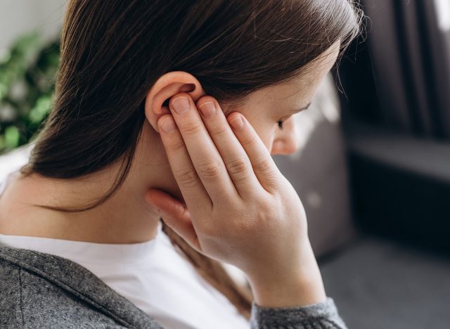 Close up of young brunette female holding painful ear, suddenly feeling strong ache. Unhealthy caucasian woman 20s suffering from painful otitis sitting on couch at home. Health problems concept