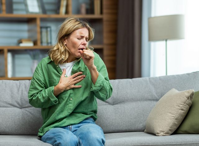 An attack of asthma, allergies at home. An elderly woman is sitting on the sofa, holding her chest and coughing, feeling severe pain, suffocating.