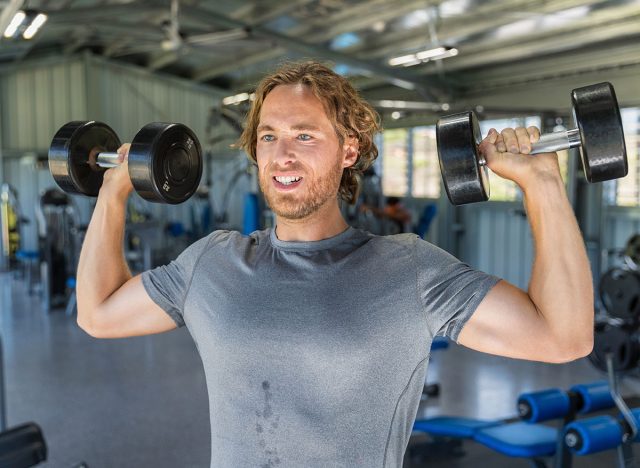 Fitness man doing military presses or standing dumbbell press exercise training in gym with free weights.