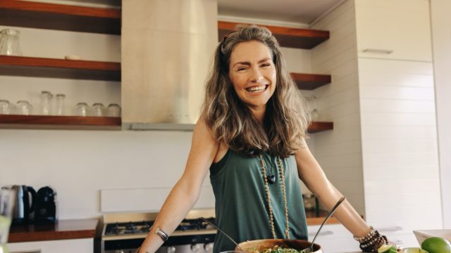 Happy vegan woman smiling at the camera while preparing a plant-based meal. Mature woman following an organic recipe in her kitchen. Healthy senior woman eating clean at home.