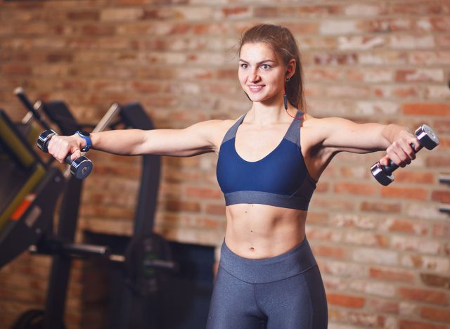 Portrait of attractive cheerful sportswoman with headphones in her ears. Sporty woman doing exercises with dumbbells for shoulders in the gym on the background of brick wall