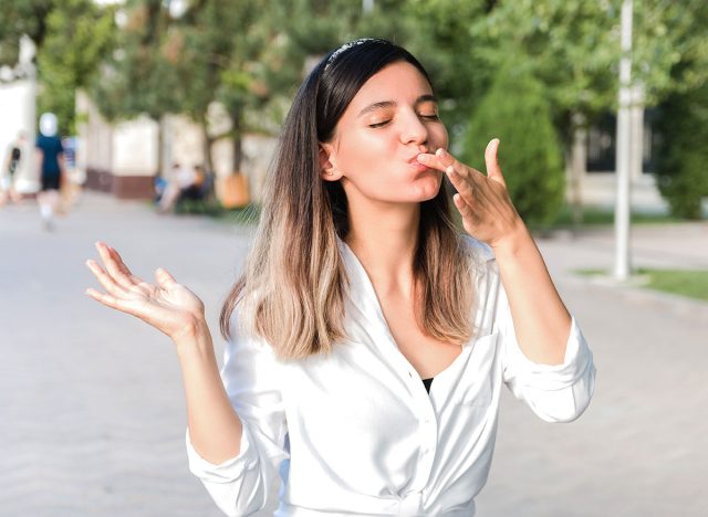 portrait of beautiful woman in white shirt and hair bezel with mouth full of food licking her fingers outdoor in city park and enjoying junk but tasty fast food while walking