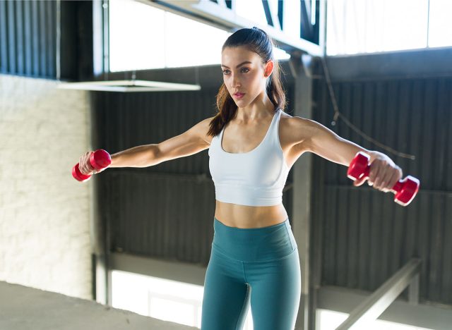 Pretty young woman with a healthy lifestyle doing side lateral raises with dumbbells indoors. Active caucasian woman exercising to stay fit and slim