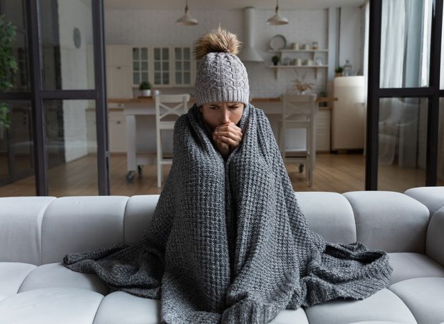Frozen. Sad latina female sit on couch at freezing cooled studio flat in warm cap and blanket shiver tremble with cold. Unhappy young lady spend time at home feel bad suffer of heating system problems