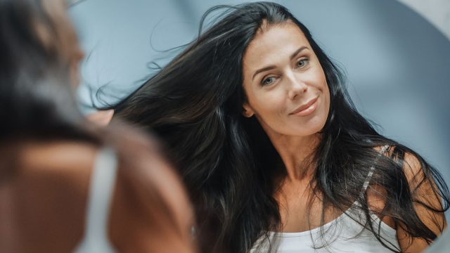Beautiful Early Middle Aged Woman Looks into Bathroom Mirror Touches Her Lush Black Hair, Admires Her Looks. Concept for Happiness, Wellbeing, Natural Beauty, Organic Skin Care Products