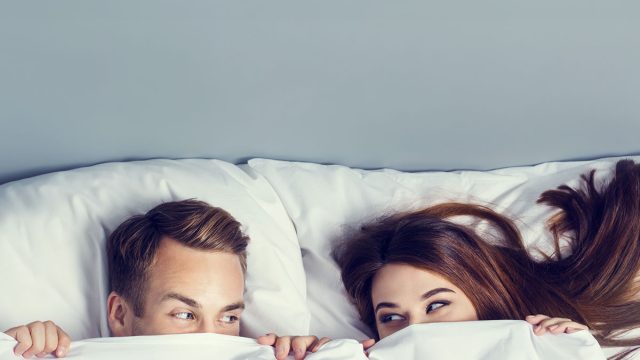 Young attractive playfull couple peeping from bedsheet on the bed at bedroom. Caucasian models in love, relationship, dating, happy people, bedtime concept shot.