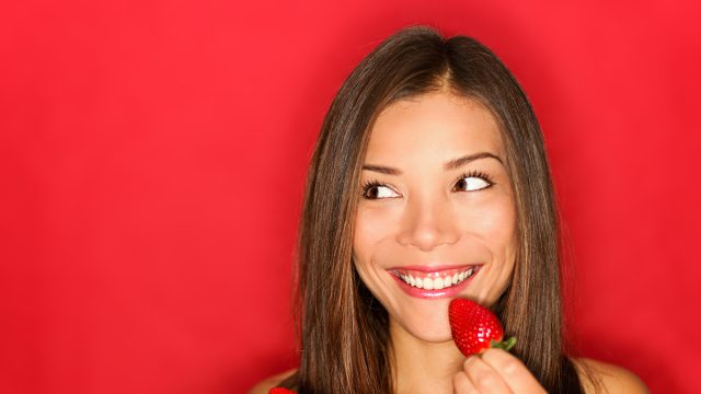 Girl eating strawberries smiling happy looking to the side on red background with copy space. Beautiful young mixed race Asian and Caucasian woman eating healthy.
