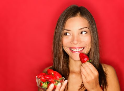 Girl eating strawberries smiling happy looking to the side on red background with copy space. Beautiful young mixed race Asian and Caucasian woman eating healthy.