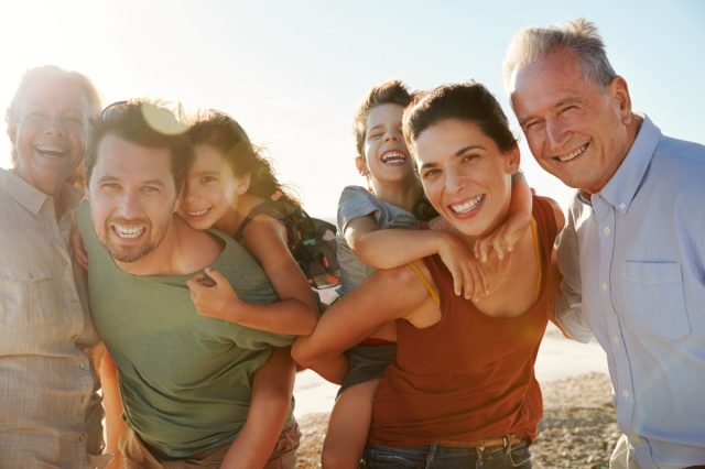 Three generation white family on a beach smiling to camera, parents piggybacking kids, close up
