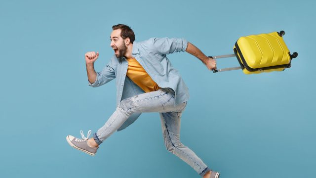Side view of funny traveler tourist man in yellow clothes isolated on blue background. Male passenger traveling abroad on weekends. Air flight journey concept. Jumping like running, hold suitcase