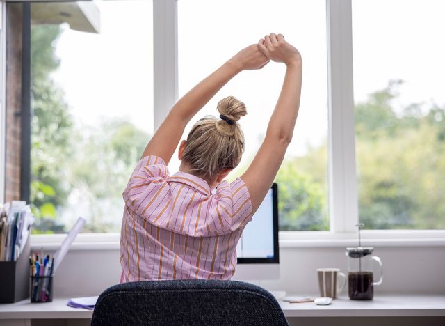 Rear View Of Woman Working From Home On Computer In Home Office Stretching At Desk