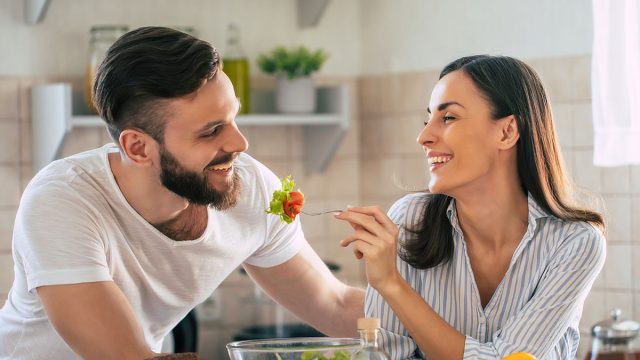 Excited smiling young couple in love making a super healthy vegan salad with many vegetables in the kitchen and man testing it from a girl's hands