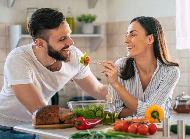 Excited smiling young couple in love making a super healthy vegan salad with many vegetables in the kitchen and man testing it from a girl's hands