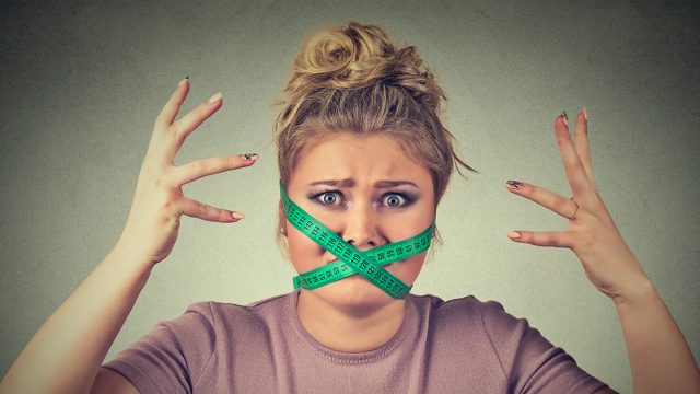 New diet restriction and stress concept. Portrait of young frustrated woman with a green measuring tape around her mouth isolated gray background. Human face expression emotion. Healthy lifestyle