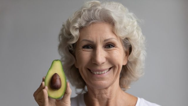 Close up head shot happy hoary senior mature retired woman looking at camera, showing avocado. Smiling healthy middle aged lady preaching vegetarian lifestyle, enjoying food vitamins, dietary concept.