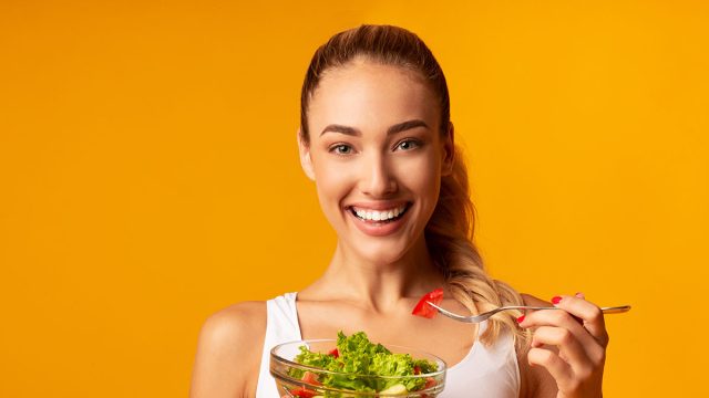 Sport And Diet. Cheerful Fitness Girl Eating Vegetable Salad Standing In Studio Over Yellow Background.