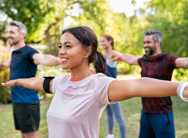 Group of multiethnic mature people stretching arms outdoor. Middle aged yoga class doing breathing exercise at park. Beautifil women and fit men doing breath exercise together with outstretched arms.
