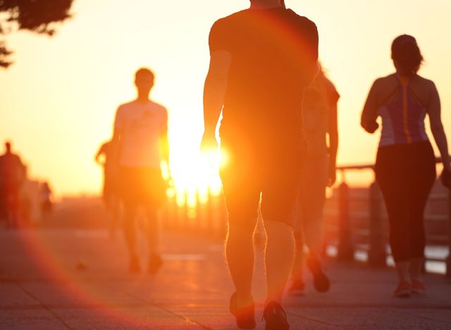 Group of unrecognized people exercise active walking on riverside boardwalk at sunset. Summer outdoor commuting fitness concept.