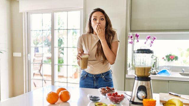 Young brunette woman drinking glass of smoothie at the kitchen covering mouth with hand, shocked and afraid for mistake.