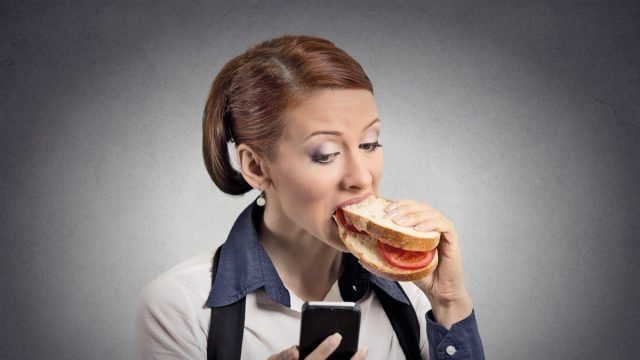 Closeup portrait young serious corporate business woman deal maker reading news message on smart mobile phone holding eating sandwich isolated grey background.
