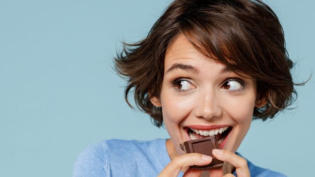 Close up young happy woman in casual sweater hold sweet pink cream donuts biting chocolate bar look camera isolated on plain pastel light blue background studio portrait. People lifestyle food concept