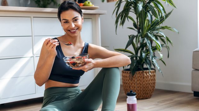 Shot of athletic woman eating a healthy bowl of muesli with fruit sitting on floor in the kitchen at home