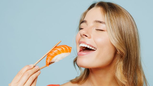Close up young woman wear orange casual clothes close eyes open mouth hold eat raw fresh sushi roll Japanese food with chopsticks isolated on plain blue background studio portrait. Lifestyle concept