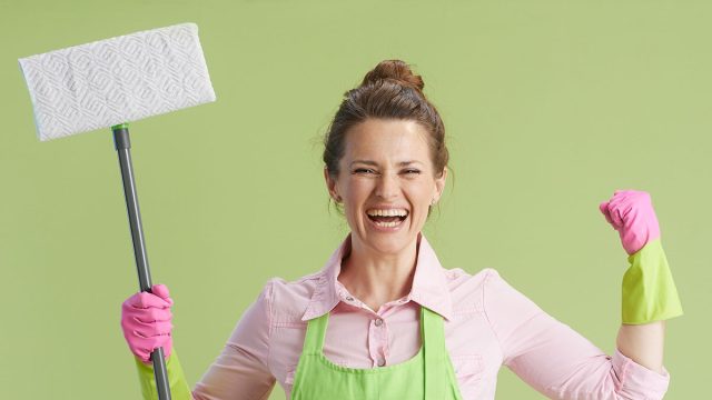 Spring cleaning. happy modern 40 years old housewife in green apron and rubber gloves against green background with mop with raised arms rejoicing.