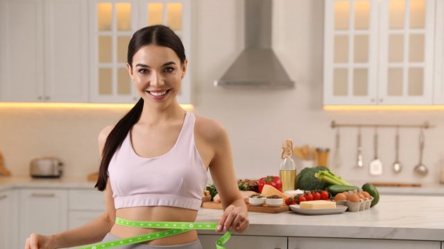 Happy woman measuring waist with tape in kitchen. Keto diet