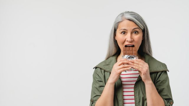 Mature middle-aged woman with grey hair eating chocolate bar.