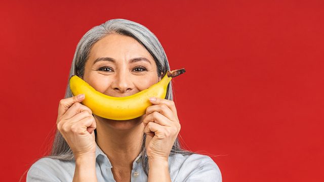 Vegan or vegetarian concept. Portrait of a beautiful elderly asian mature aged woman holding banana fruit, smiling, isolated over red background.