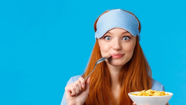 Morning, lifestyle and women concept. Pretty cute redhead, foxy girl with long red hair waking-up early, prepare breakfast, biting spoon and smiling eating cereals, standing blue background.