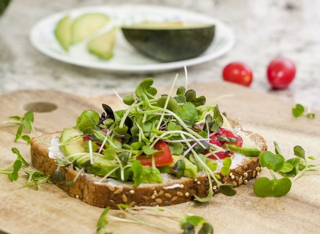 A delicious open-faced sandwich is displayed on a wooden board, featuring a generous layer of avocado, sliced tomatoes, and an abundance of vibrant microgreens