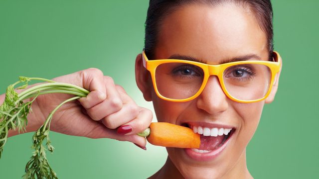 Young woman wearing glasses while eating fresh carrot. Closeup portrait of a young woman wearing glasses while eating carrot against green background.