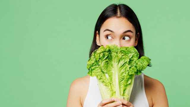 Young woman wear white clothes hold cover mouth with bunch of fresh greens lettuce leaves look aside isolated on plain pastel light green background. Proper nutrition healthy fast food choice concept