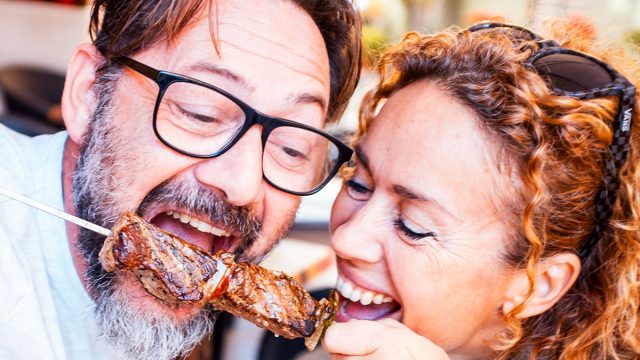 Happy adult couple have fun together eating meat cooked on a spit in touristic restaurant. Man and woman enjoy time eating meal from same fork posing for a picture. Happiness relationship food concept