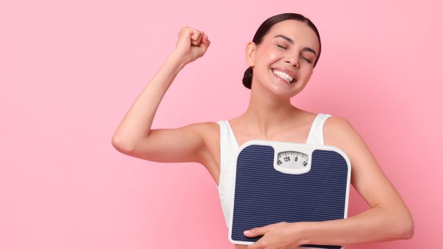 Diet and weight loss concept. Happy young woman with floor scale on pink background. Space for text