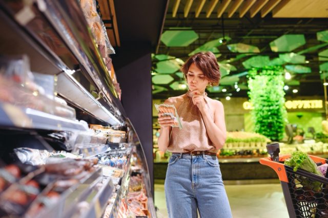 Young woman in casual clothes at supermarket store looking at meat sausage and read ingredients