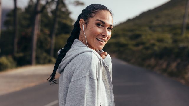Rear view of young woman walking on empty road and looking over her shoulder. Sporty woman on country road with earphones on.