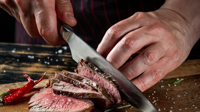Cooking juicy beef steak by chef hands on dark black background with copy space for text menu or recipe.